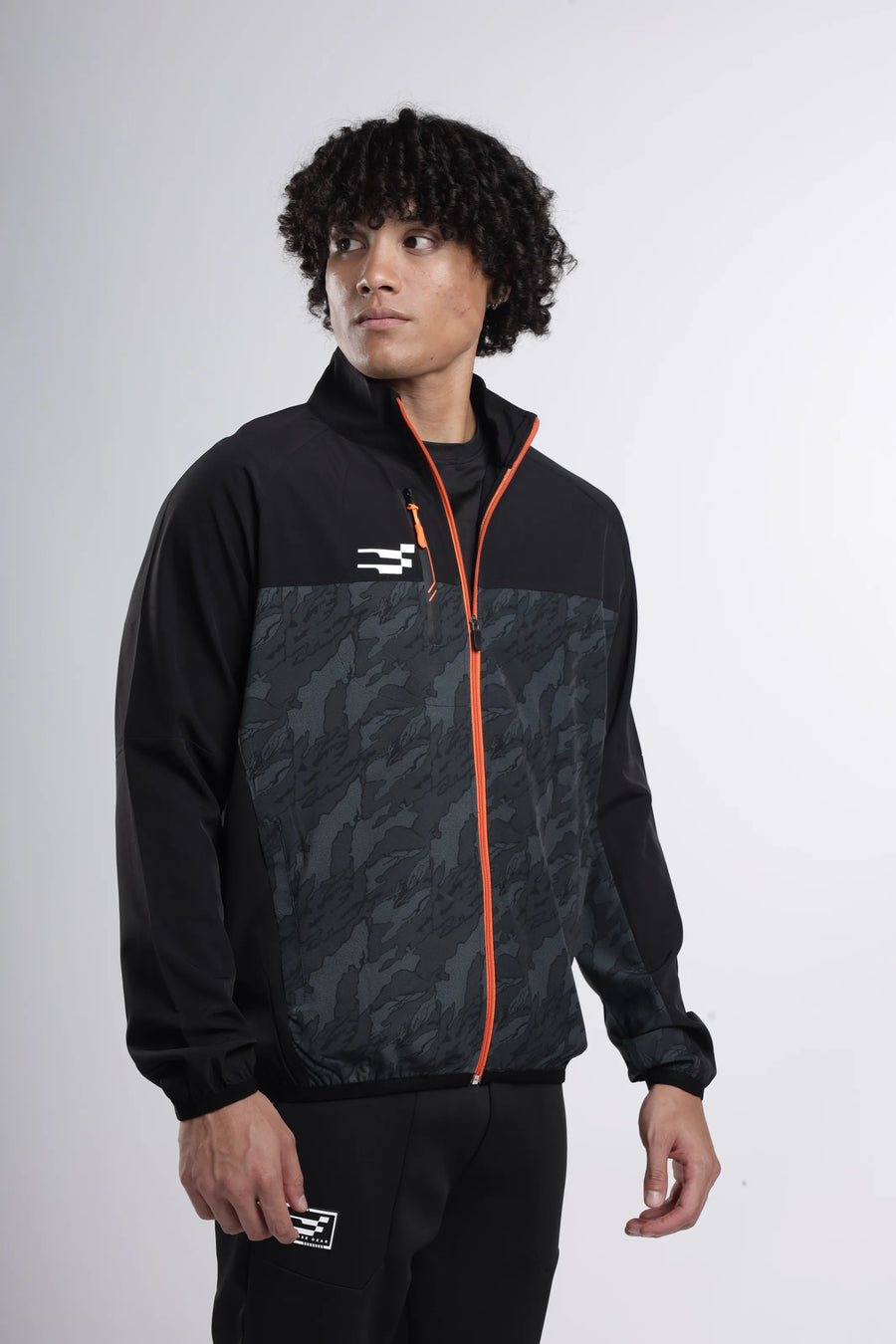 Quantum Performance Track-Top in black, three-quarter view showing the camo panel, orange zip and chest logo